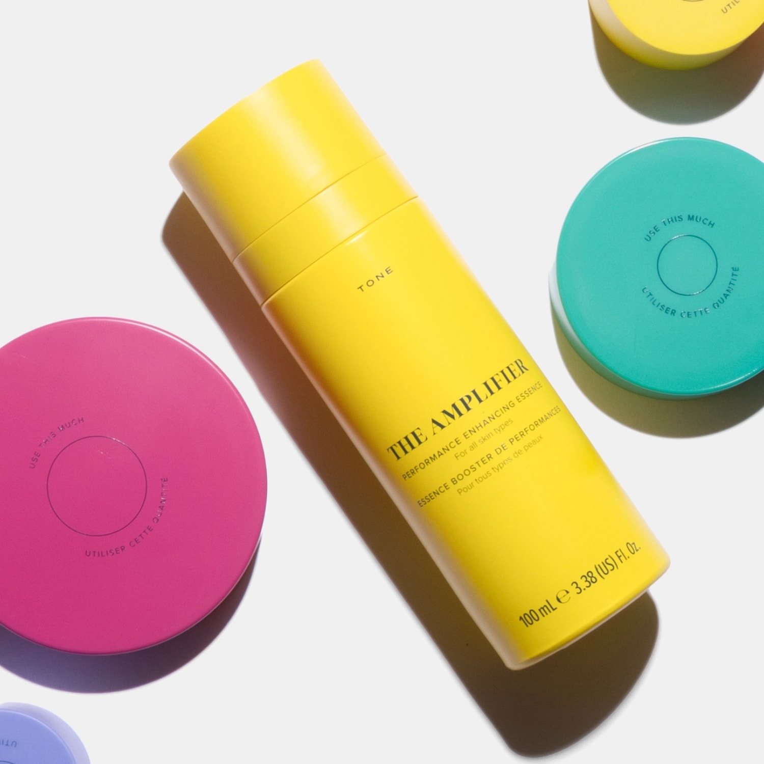 The Amplifier Performance Enhancing Essence for all skin types comes in a vibrant yellow bottle, containing 100 ml or 3.38 US fl. oz. of product, surrounded by colorful circular containers in pink, teal, and yellow, each marked with "USE THIS MUCH" and "UTILISER CETTE QUANTITÉ".