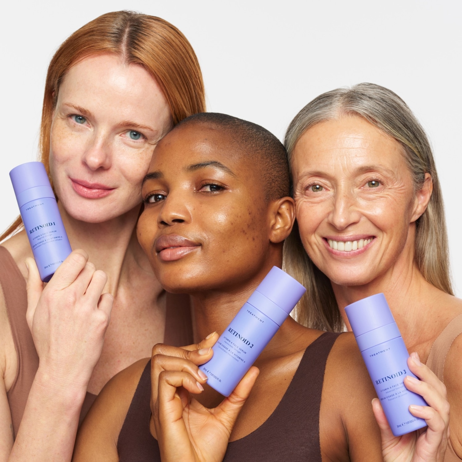 Three women with diverse skin tones and hair colors, wearing brown tops, hold up purple bottles of "Retinoid 1," "Retinoid 2," and "Retinoid 3" Vitamin A Face Serum, showcasing the product range.