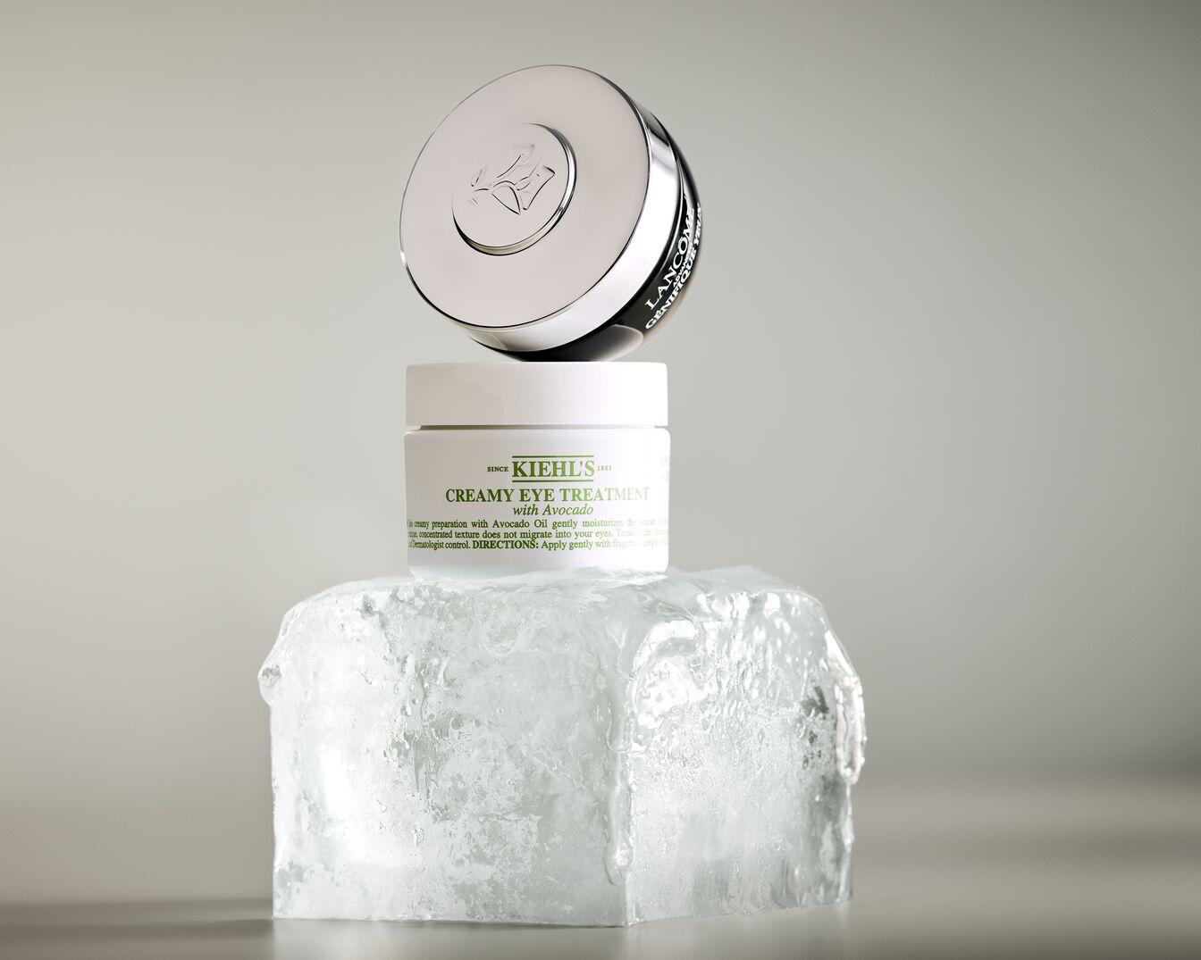 Un pot blanc de crème contour des yeux Kiehl's à l'avocat est posé sur un bloc de glace, avec un couvercle argenté de Lancôme Génifique Yeux en équilibre au-dessus.