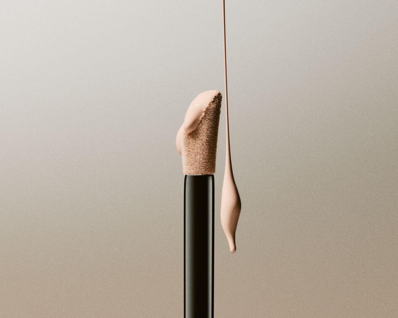 Un applicateur de correcteur noir avec une pointe floquée beige est visible, avec une goutte de correcteur beige clair qui coule de l'applicateur et une autre goutte suspendue en dessous, le tout sur un fond beige uni.