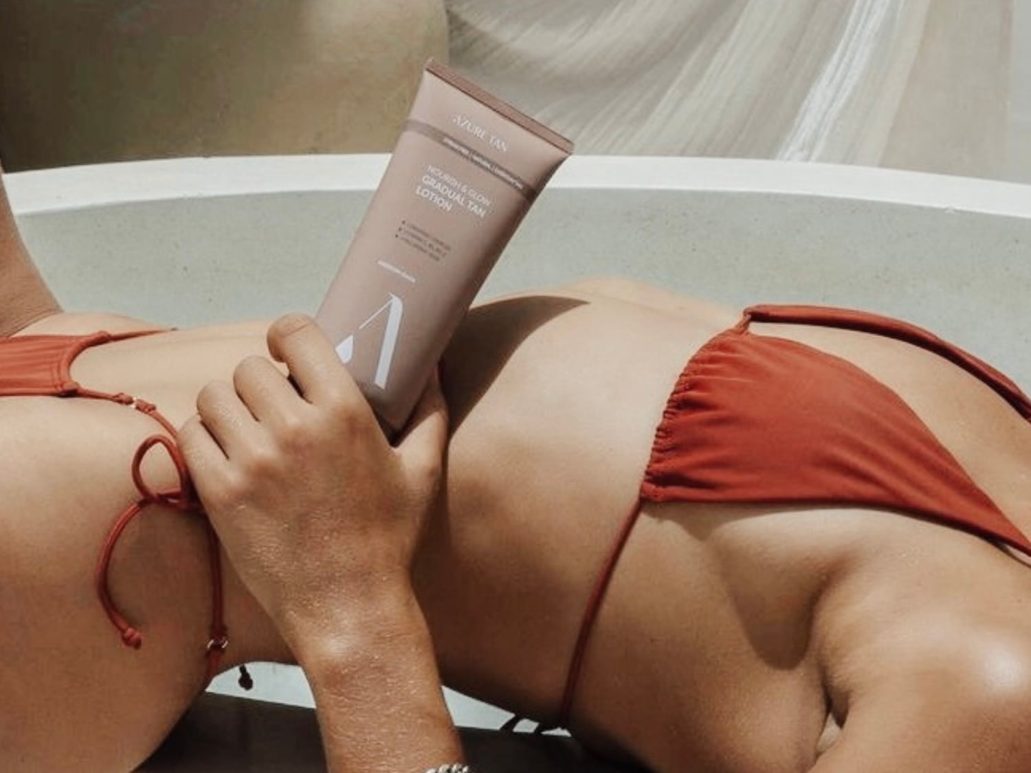 Osoba w pomarańczowym bikini leży na plecach, trzymając w dłoni beżową tubkę balsamu do opalania Azure Tan Nourish & Glow Gradual Tan Lotion.