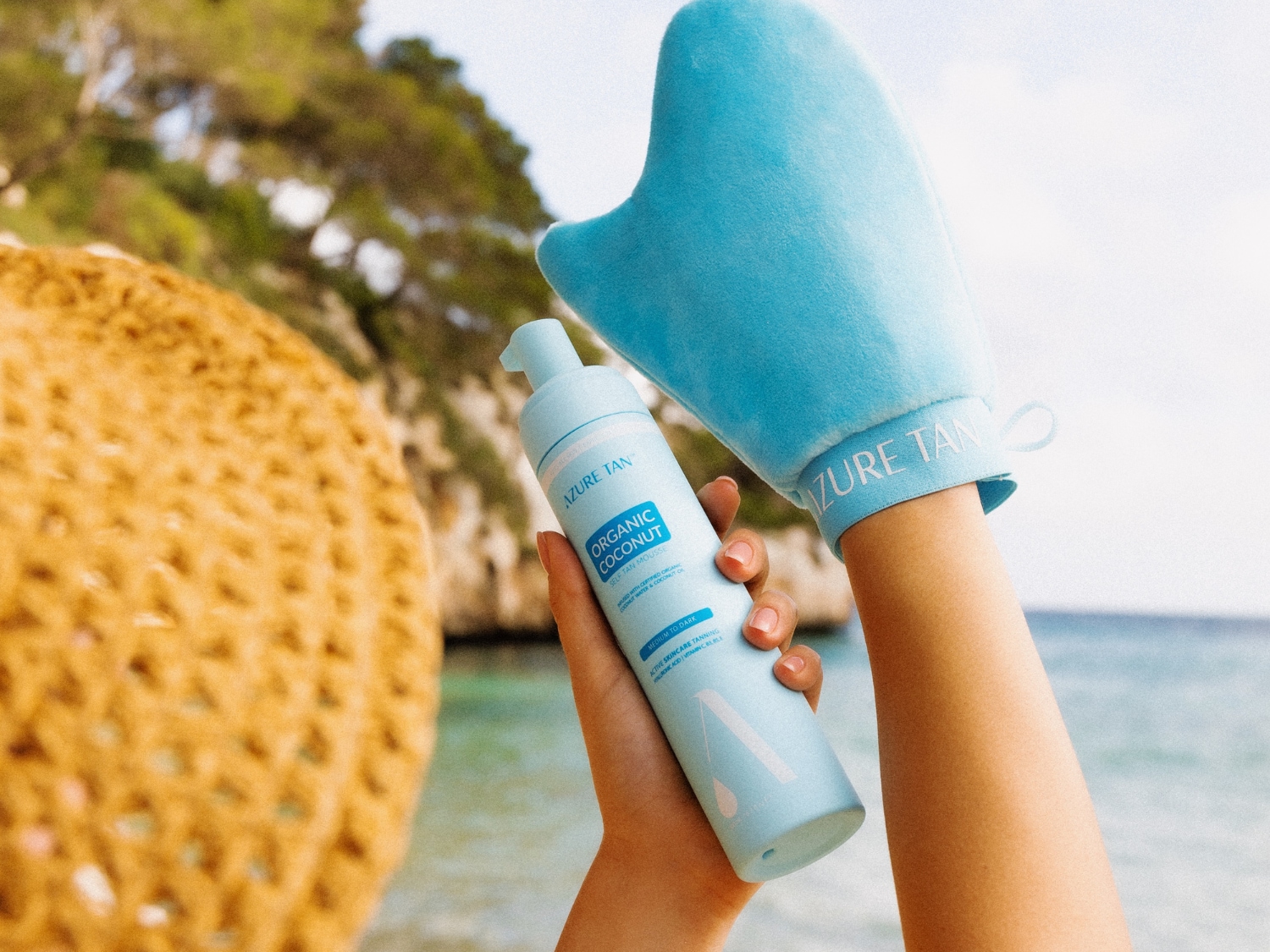 Dłoń trzyma jasnoniebieską butelkę samoopalacza w piance Azure Tan Organic Coconut Self Tan Mousse oraz jasnoniebieską rękawicę do aplikacji z napisem "Azure Tan", na tle plaży i morza.