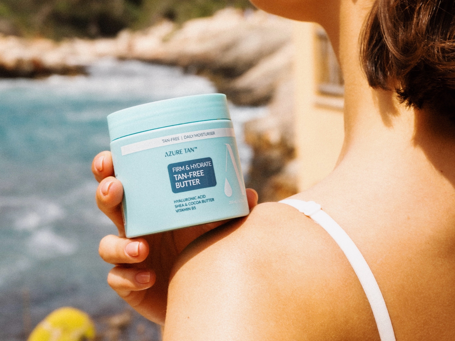 Krem Azure Tan Firm & Hydrate Tan-Free Butter w jasnoniebieskim słoiczku, wzbogacony kwasem hialuronowym, masłem shea i kakaowym oraz witaminą B5, jest trzymany przez kobietę na tle błękitnego morza.
