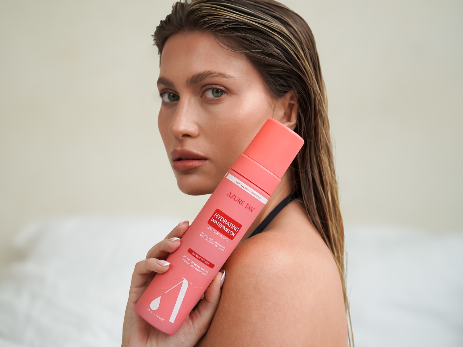 Modelka trzyma w dłoni różową butelkę samoopalacza Azure Tan Hydrating Watermelon Self Tan Mousse, który jest odpowiedni dla każdego rodzaju skóry i zapewnia ekspresową opaleniznę od średniej do ciemnej.