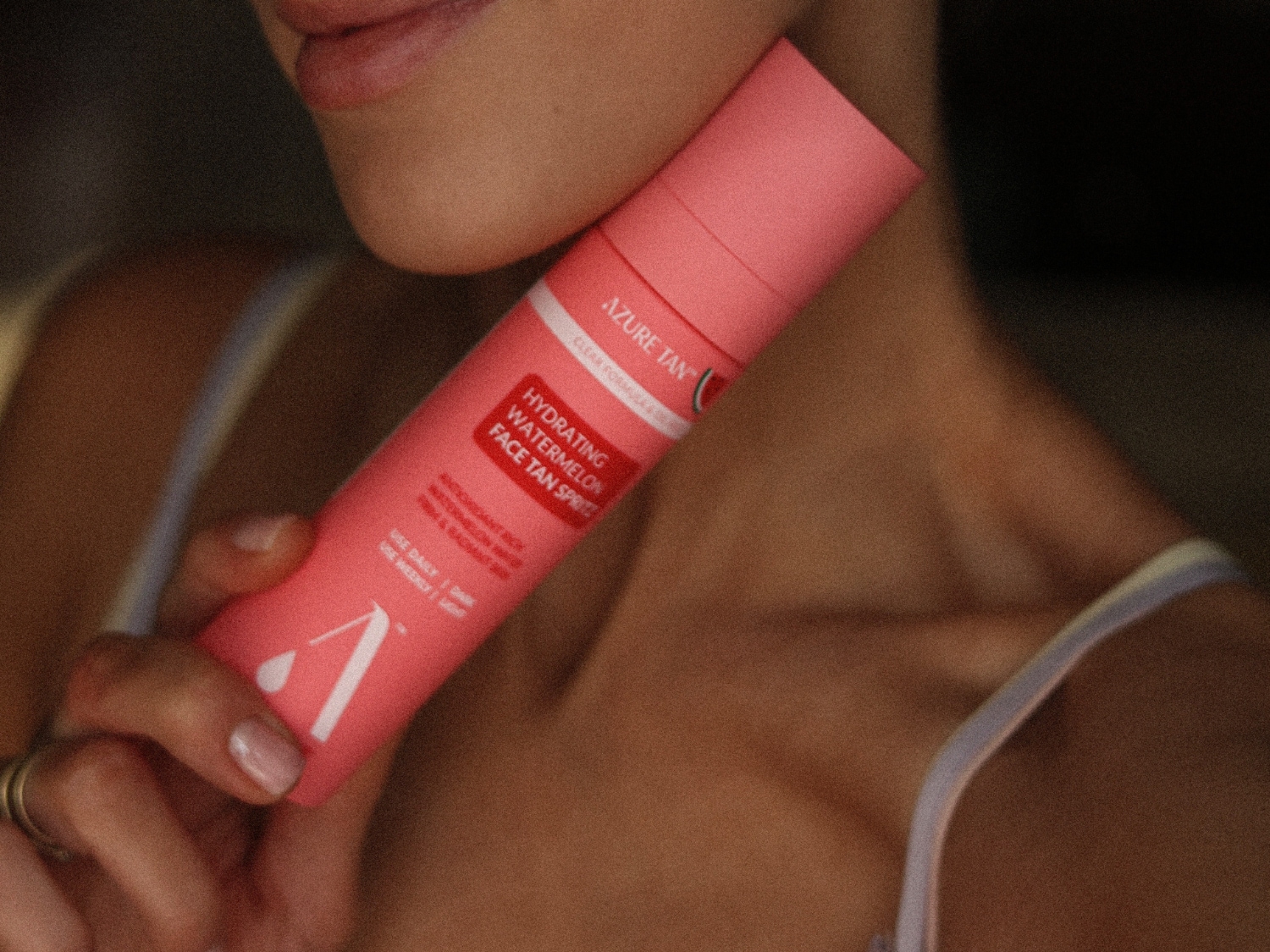 Kobieta trzyma w dłoni różowy spray do opalania twarzy Azure Tan Hydrating Watermelon Face Tan Spritz, z widocznym napisem „Clear Formula” i „Use Daily / Dark, Use Weekly / Light”.