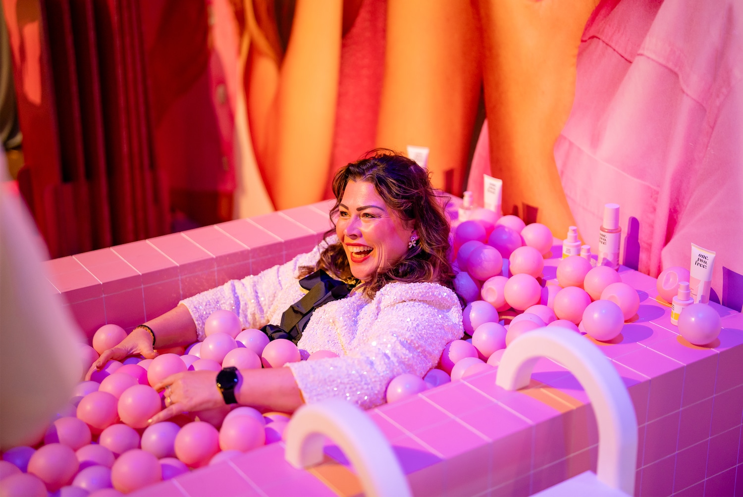 Een lachende vrouw met donker haar en een glinsterend wit jasje ligt in een roze ballenbak, omringd door roze ballen en enkele huidverzorgingsproducten in roze verpakkingen.