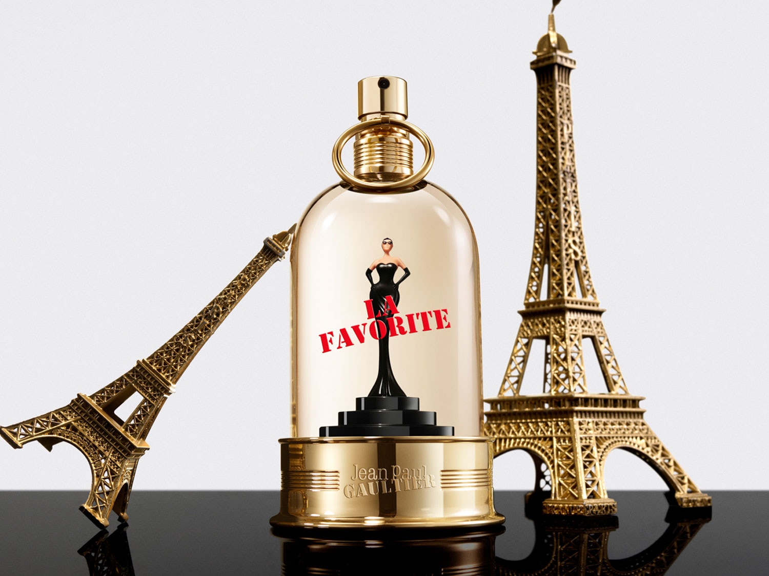 Le parfum Jean Paul Gaultier La Favorite est présenté dans un flacon doré avec une figurine de femme en robe noire à l'intérieur, flanqué de deux répliques dorées de la Tour Eiffel.