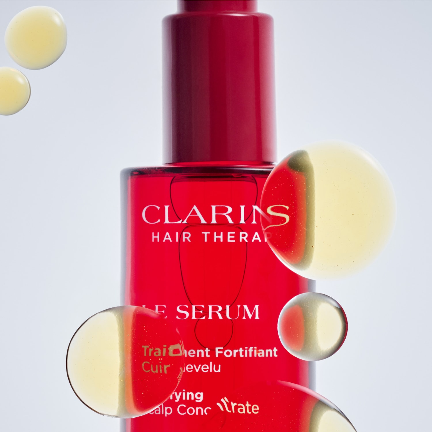 A red bottle of Clarins Hair Therapy serum with a red cap is shown, with several golden oil droplets suspended around it, highlighting the product's texture and ingredients.