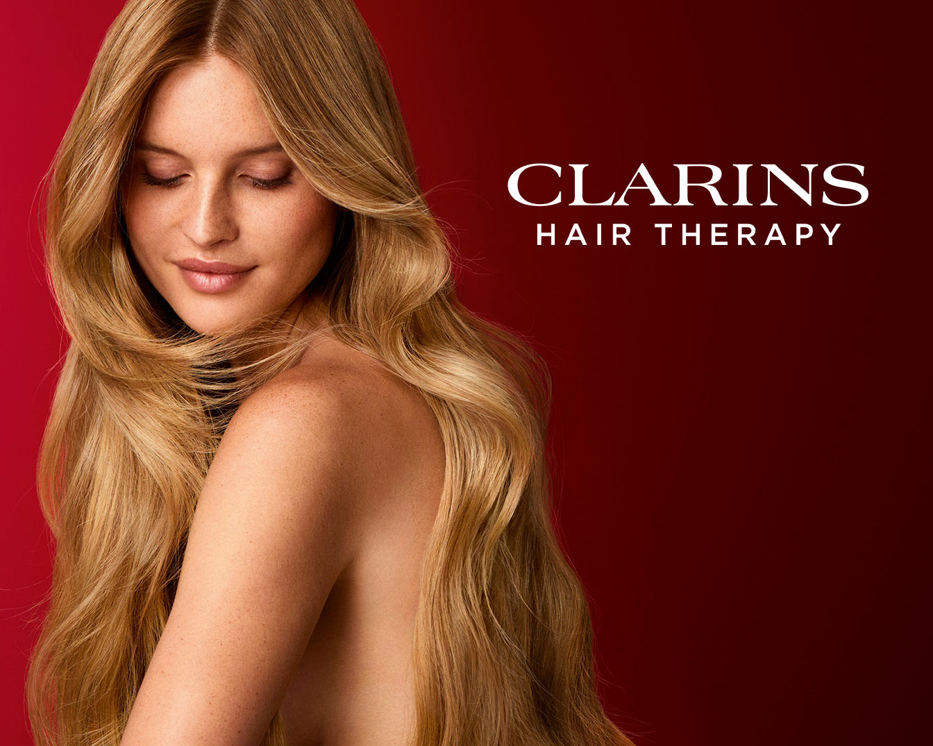 A woman with long, wavy blonde hair and freckles on her shoulders and face, looks down with closed eyes, promoting Clarins Hair Therapy against a deep red background.