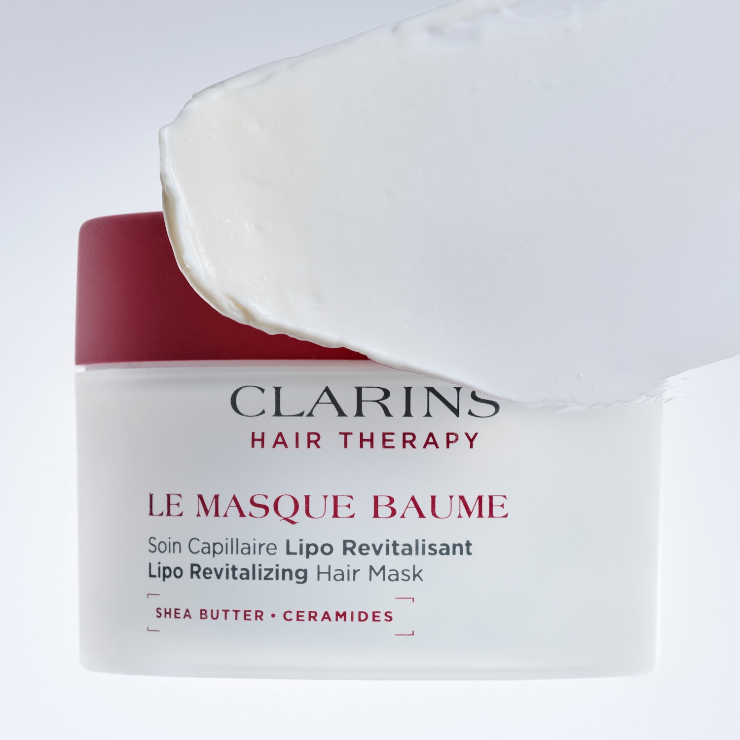 Clarins Hair Therapy Le Masque Baume Lipo Revitalizing Hair Mask, in a white jar with a red lid, features a rich, white cream texture, enriched with Shea Butter and Ceramides for revitalized hair.