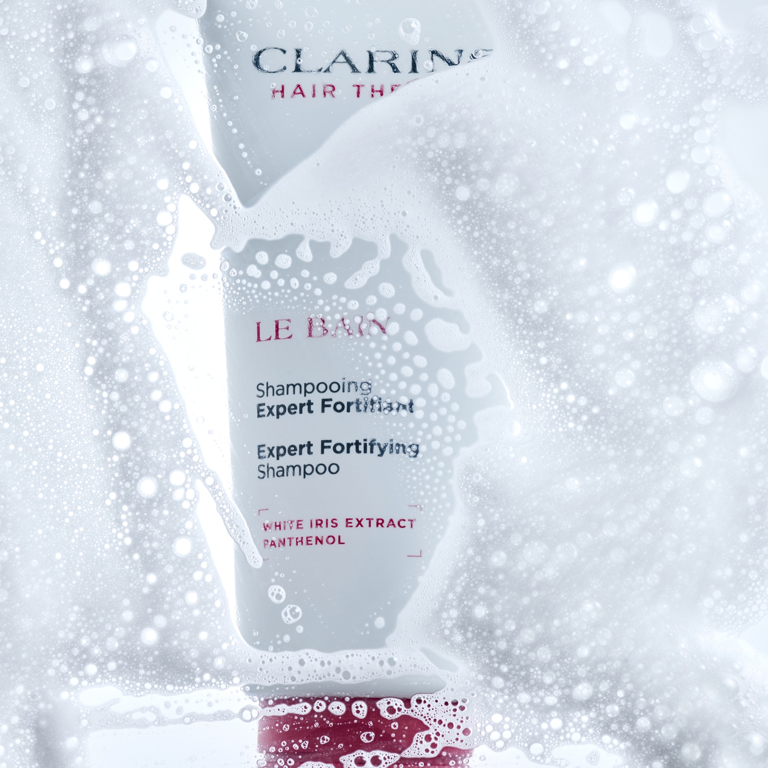 A white bottle of Clarins Expert Fortifying Shampoo, enriched with White Iris Extract and Panthenol, is covered in white foam, highlighting its cleansing properties.