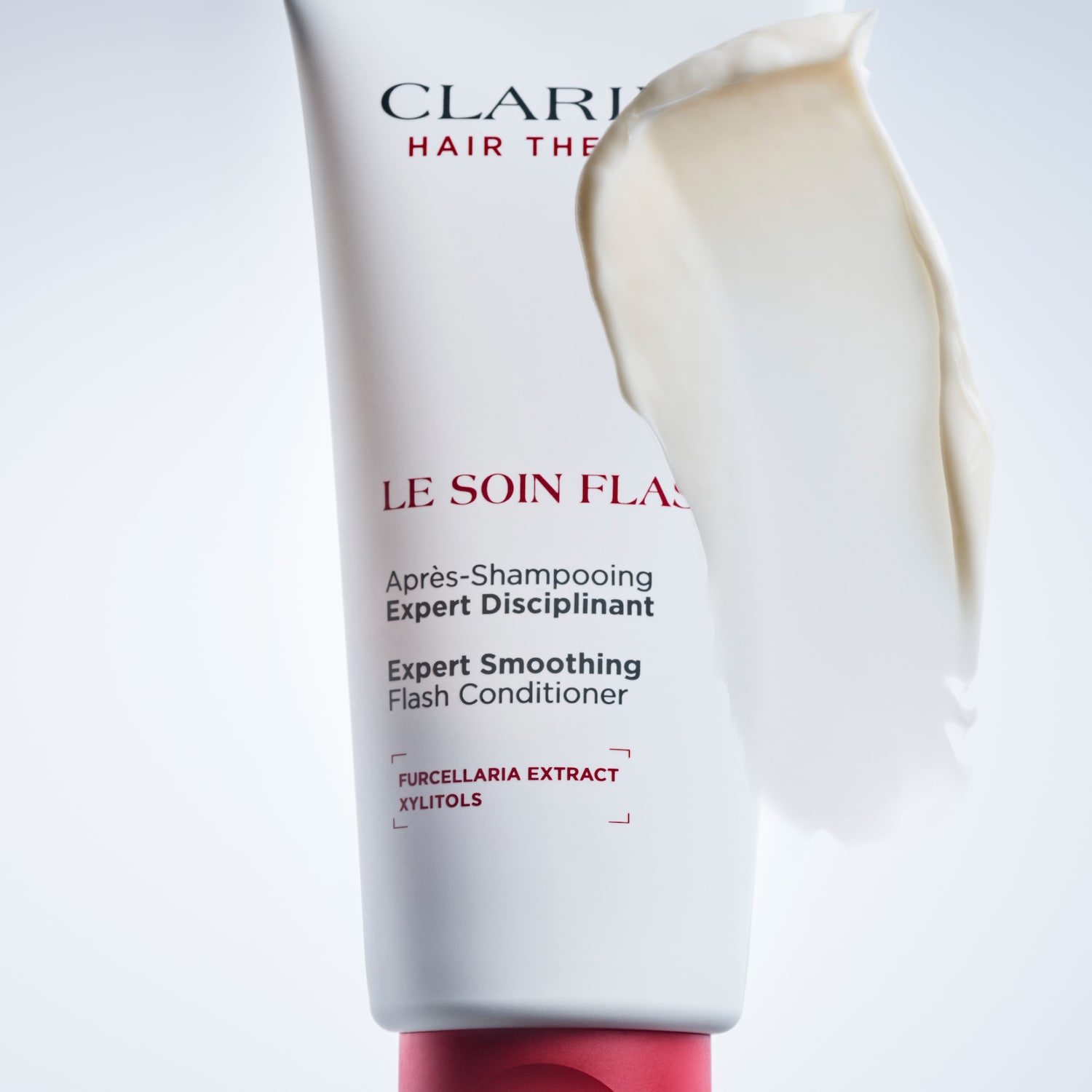 Clarins Expert Smoothing Flash Conditioner in a white tube with a red cap, featuring a creamy white product swatch on the side, enriched with Furcellaria Extract and Xylitols for expert hair care.