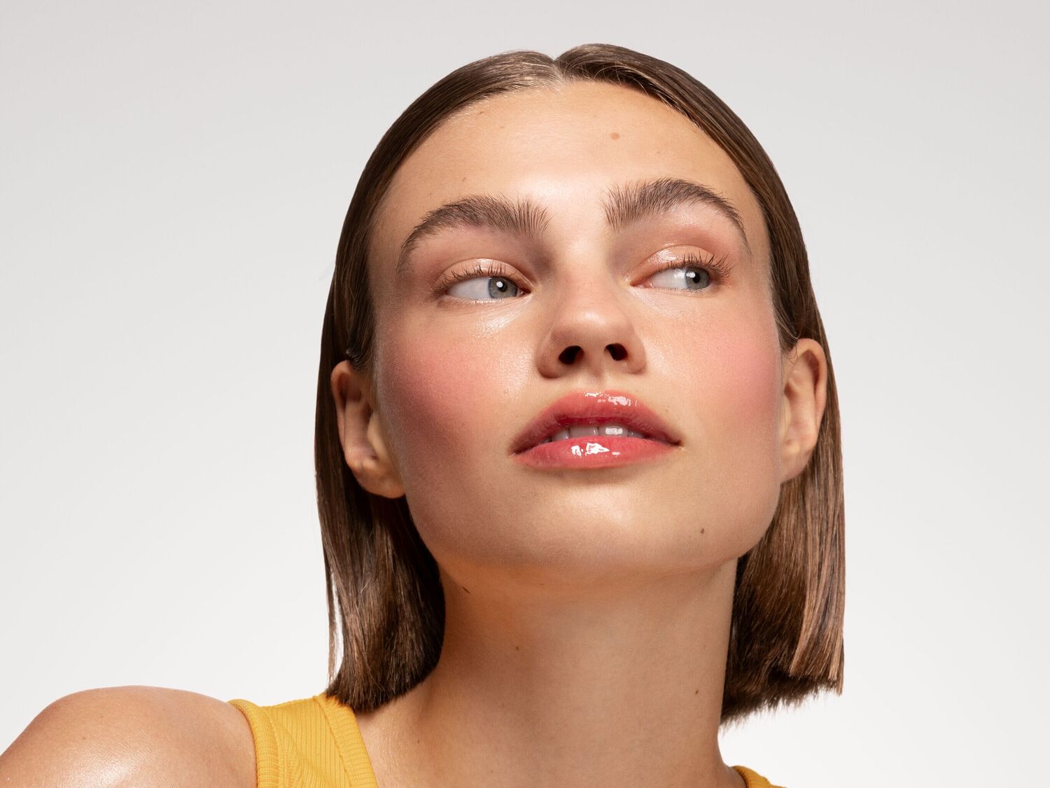 Une femme aux cheveux courts et bruns, vêtue d'un haut jaune, présente un maquillage frais avec un blush rosé sur les joues et un gloss brillant sur les lèvres, regardant vers la droite.