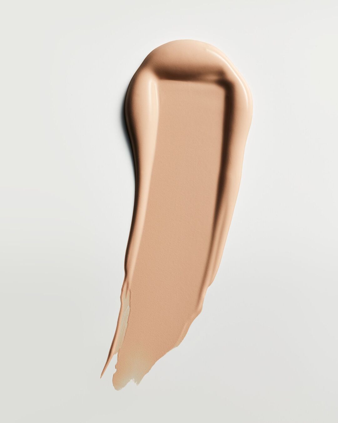 Un échantillon de fond de teint liquide beige clair est étalé en diagonale sur un fond blanc, révélant sa texture lisse et crémeuse.