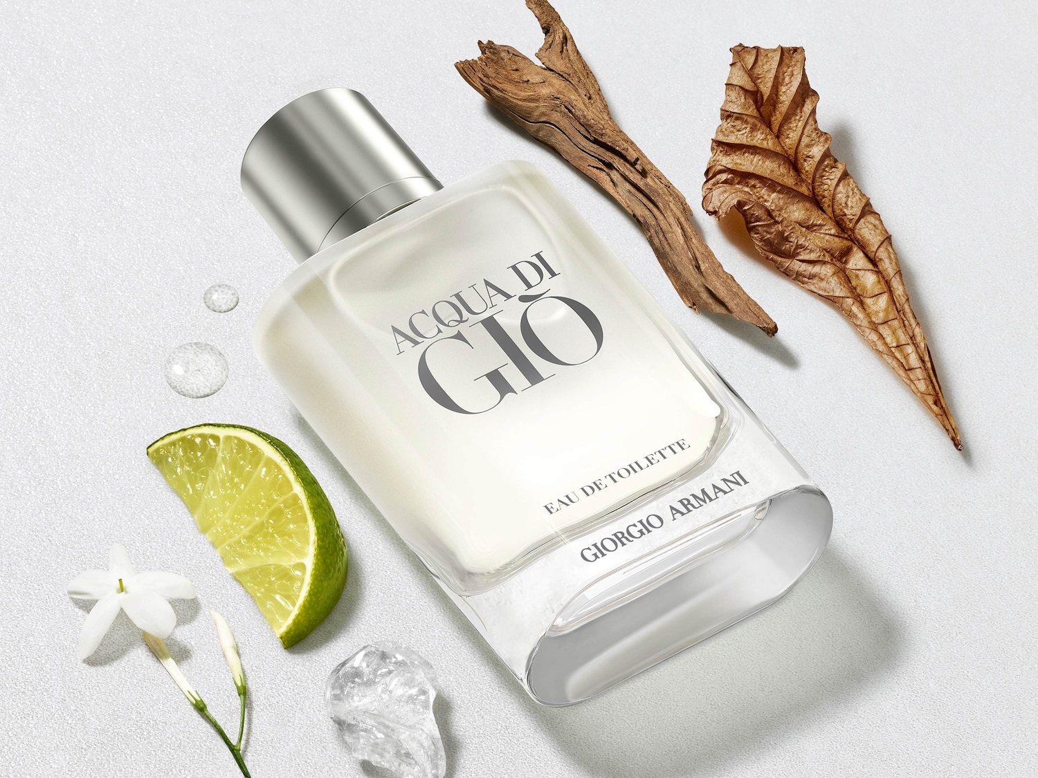 Ein Flakon von Acqua di Gio Eau de Toilette von Giorgio Armani, umgeben von einer Limettenscheibe, Jasminblüten, Wassertropfen, einem Stück Treibholz und einem getrockneten Blatt, präsentiert sich auf einem strukturierten weißen Hintergrund.