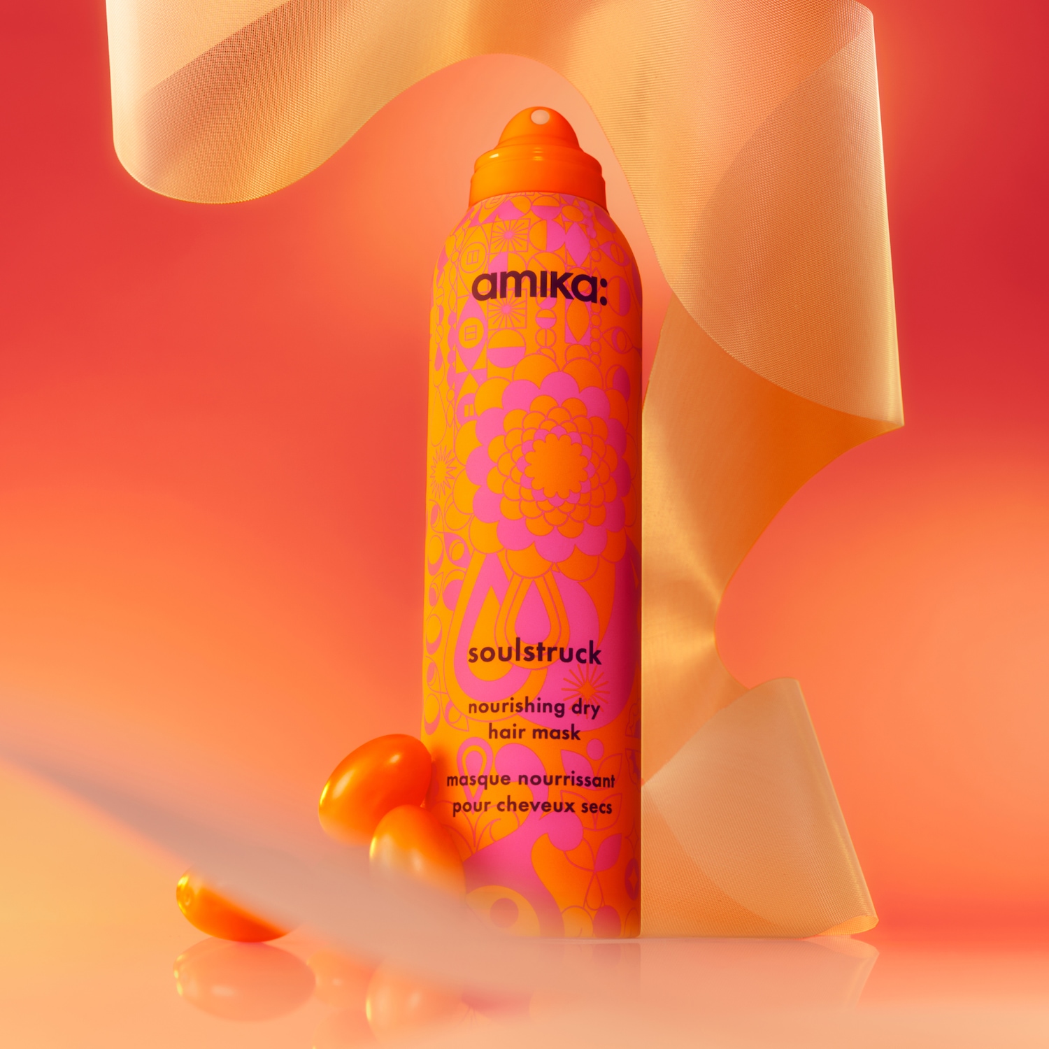 Die Amika Soulstruck Nourishing Dry Hair Mask in einer leuchtend orangefarbenen Flasche mit pinken und gelben Blumenmustern, umgeben von einem fließenden, transparenten orangefarbenen Band und drei kleinen orangefarbenen Früchten, die auf einem glänzenden, orange-roten Hintergrund liegen.