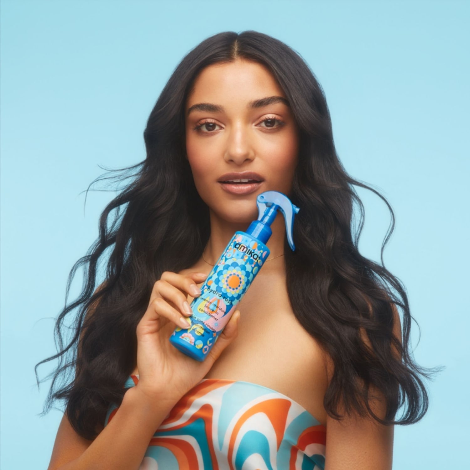Eine Frau mit langen, dunklen Haaren hält eine blaue Sprühflasche von Amika Hydro Rush Intense Moisture Leave-In Conditioner mit einem bunten Blumenmuster in der Hand.