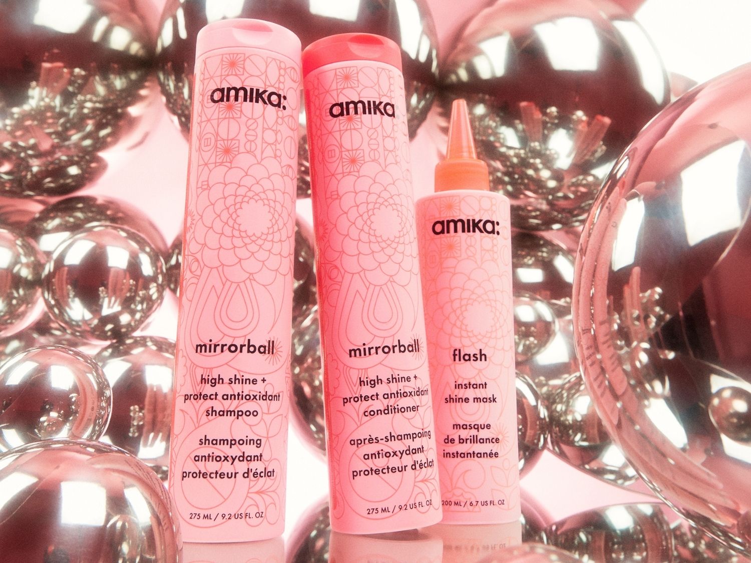 Drei Amika-Produkte, darunter Shampoo, Spülung und eine glänzende Maske, stehen aufrecht vor einem Hintergrund aus glänzenden, rosafarbenen Kugeln, die einen festlichen Glanz verleihen.