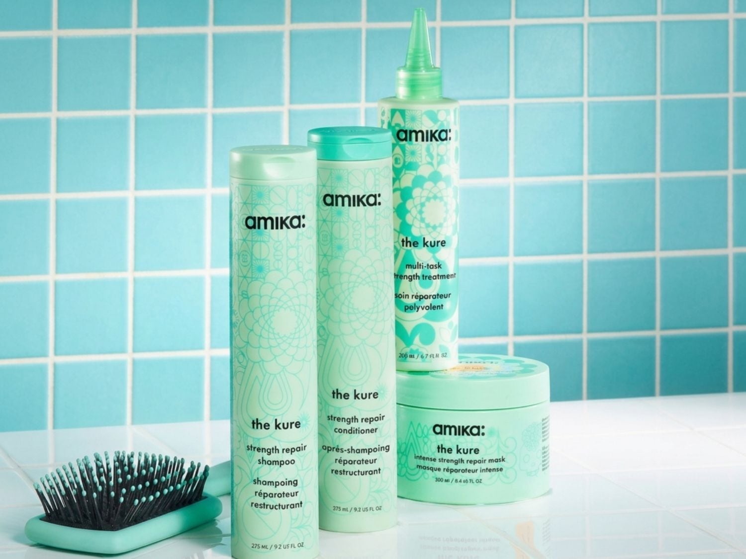 Das Bild zeigt eine Reihe von Amika The Kure Haarpflegeprodukten in mintgrünen Flaschen und Tiegeln, darunter Shampoo, Spülung, eine Multitasking-Stärkungsbehandlung und eine intensive Reparaturmaske, zusammen mit einer passenden Haarbürste, die alle auf einer weißen Oberfläche vor einem hellblauen Fliesenmuster stehen.