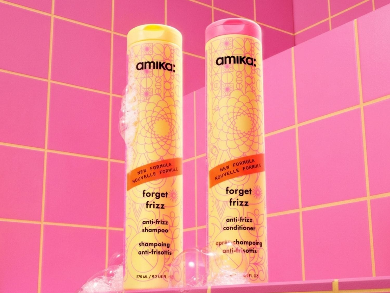 Zwei Amika "Forget Frizz" Anti-Frizz Shampoo und Conditioner Flaschen stehen vor einem pinken Fliesenmuster, umgeben von Seifenblasen.
