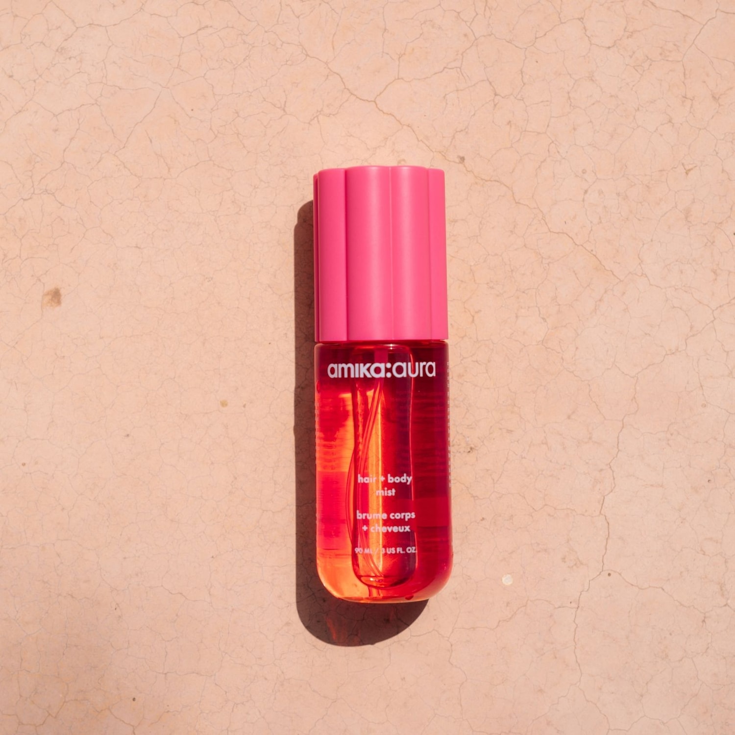 Das amika:aura Haar- und Körperspray in einer transparenten Flasche mit leuchtend pinkem Deckel und roter Flüssigkeit, die sich unten orange färbt, steht auf einem hellbraunen, rissigen Untergrund.