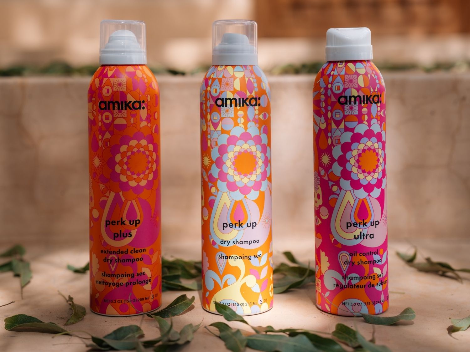 Drei bunte Flaschen Amika Trockenshampoo stehen auf einem hellbraunen Untergrund, umgeben von grünen Blättern, die die verschiedenen Varianten Perk Up Plus, Perk Up und Perk Up Ultra präsentieren.