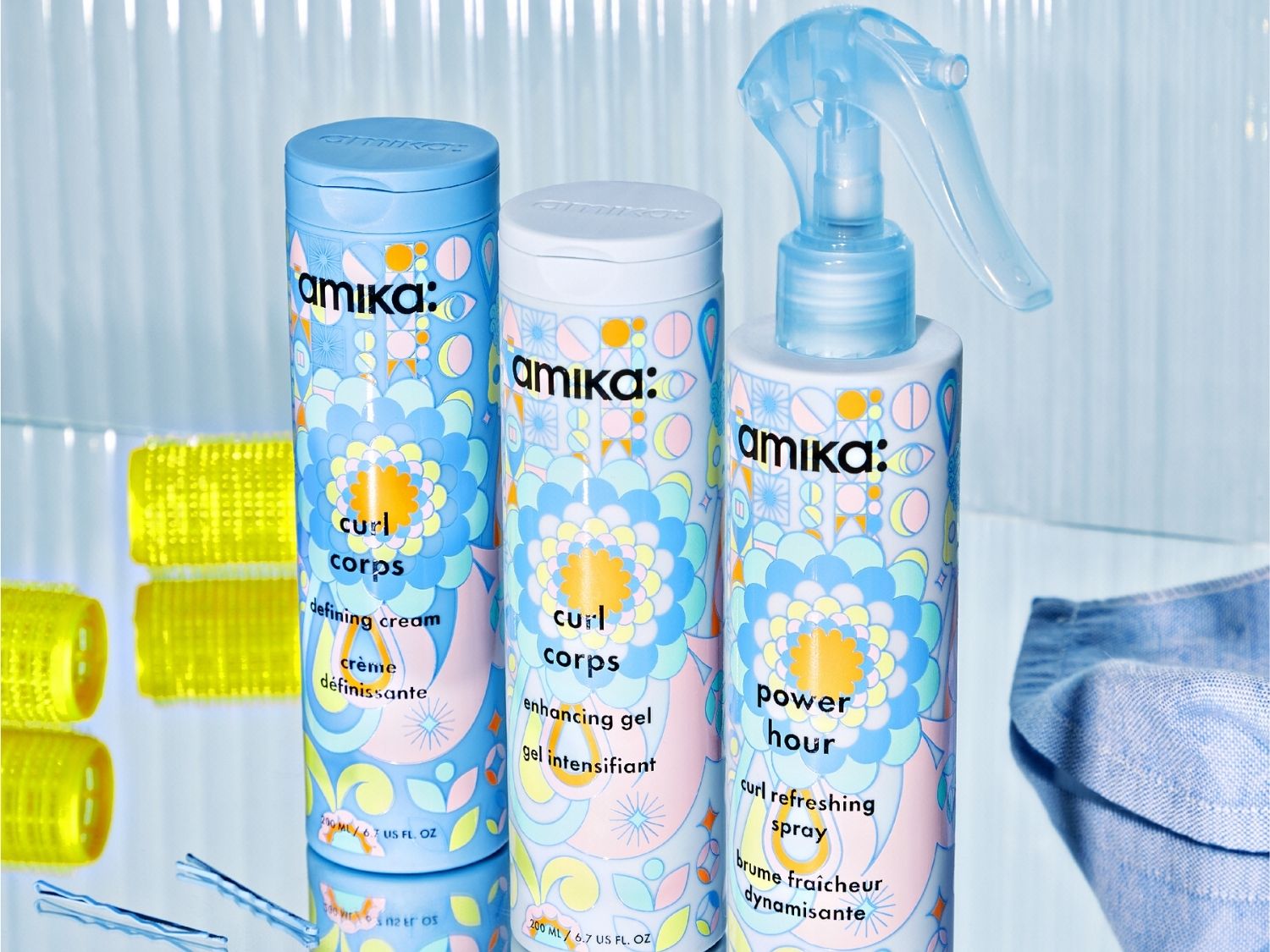 Drei Amika-Haarpflegeprodukte in hellblauen Flaschen mit bunten Blumenmustern stehen auf einer spiegelnden Oberfläche, darunter eine "Curl Corps Defining Cream", ein "Curl Corps Enhancing Gel" und ein "Power Hour Curl Refreshing Spray".