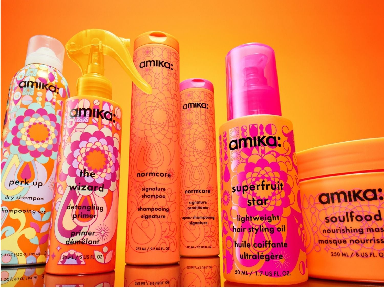 Eine Reihe von Amika-Haarpflegeprodukten in leuchtend orangefarbenen Flaschen mit bunten Blumenmustern, darunter Trockenshampoo, Entwirrungsgrundierung, Shampoo, Spülung, leichtes Haarstylingöl und eine nährende Maske.