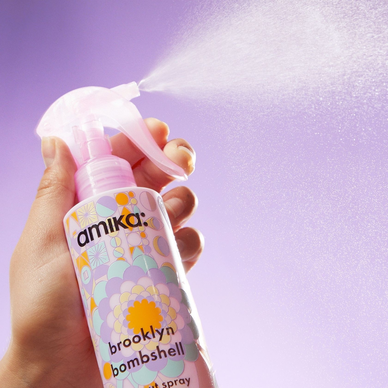 Eine Hand hält eine pinkfarbene Sprühflasche von Amika Brooklyn Bombshell, die ein feines Spray vor einem lila Hintergrund abgibt.