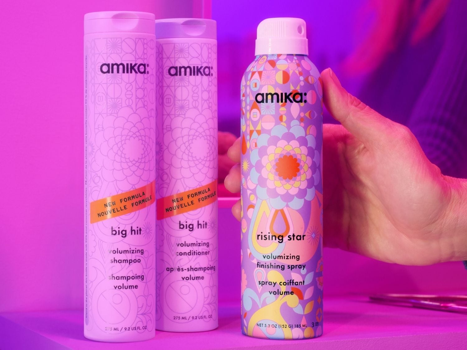 Drei Amika-Haarpflegeprodukte stehen vor einem violetten Hintergrund, darunter zwei "Big Hit" Volumizing Shampoo und Conditioner in hellvioletten Flaschen mit Blumenmuster und ein "Rising Star" Volumizing Finishing Spray in einer bunten Dose, die von einer Hand gehalten wird.