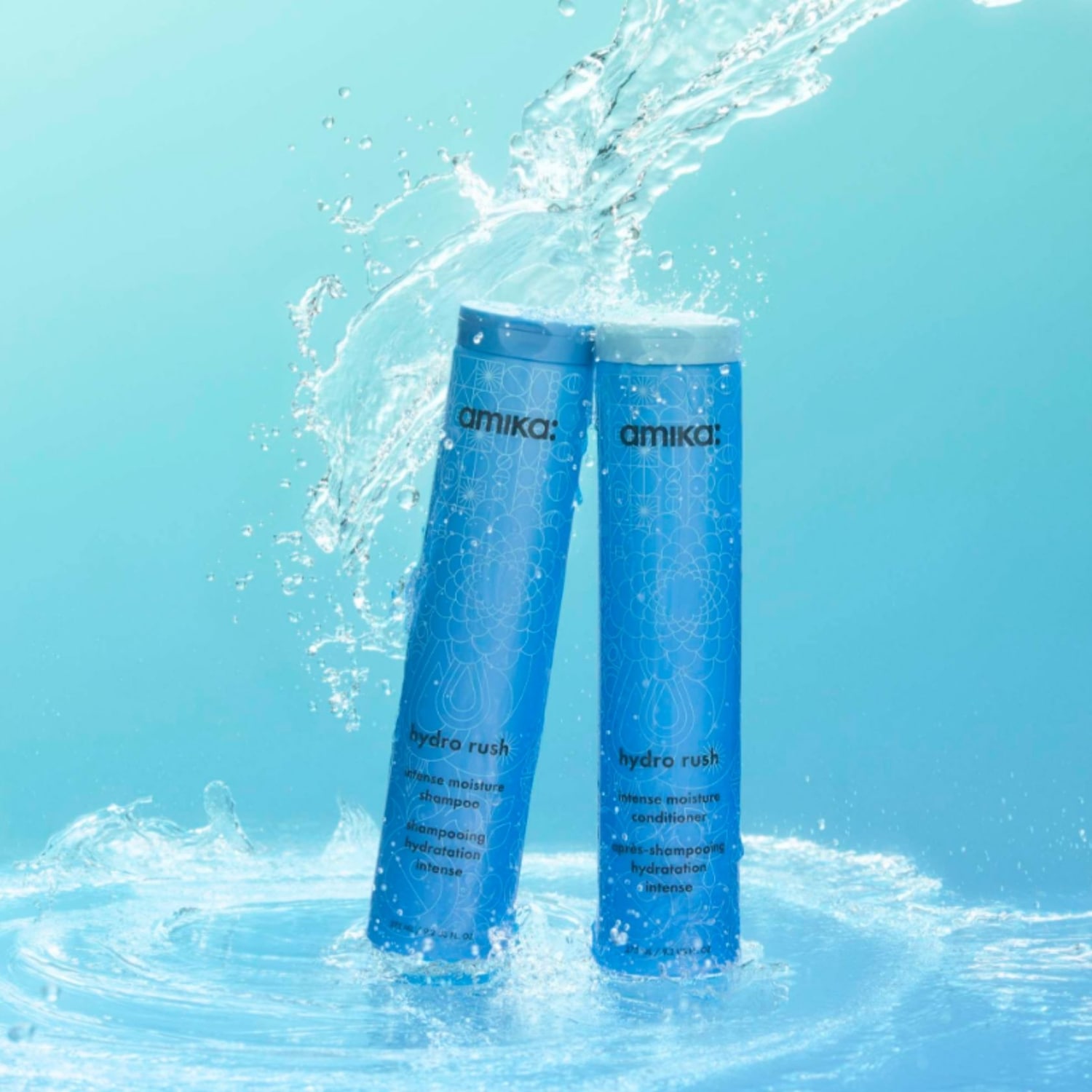 Zwei blaue Flaschen von Amika Hydro Rush Intense Moisture Shampoo und Conditioner stehen in spritzendem Wasser vor einem hellblauen Hintergrund.