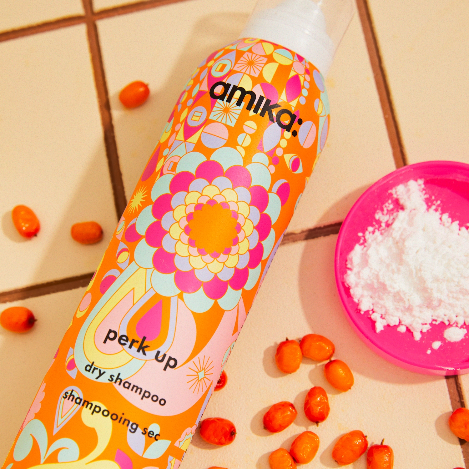 Das Amika Perk Up Trockenshampoo in einer farbenfrohen, gemusterten orangefarbenen Flasche mit weißem Sprühkopf steht auf einem beigefarbenen Fliesenboden, umgeben von kleinen orangefarbenen Beeren und einer pinken Schale mit weißem Puder.