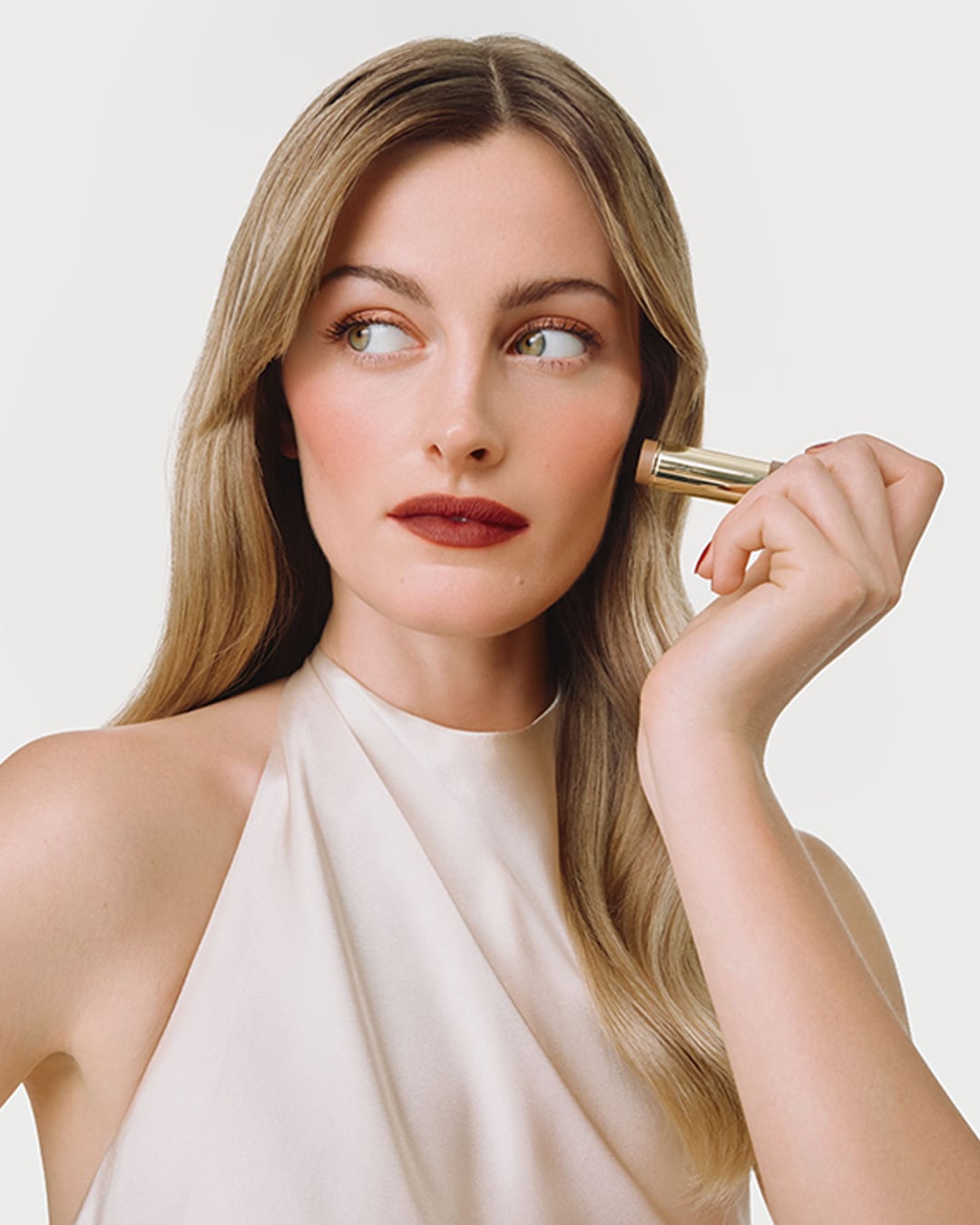 Eine Frau mit langen blonden Haaren, die ein cremefarbenes Oberteil trägt, trägt einen goldenen Make-up-Stift auf ihre Wange auf, um ihre Gesichtszüge zu betonen.