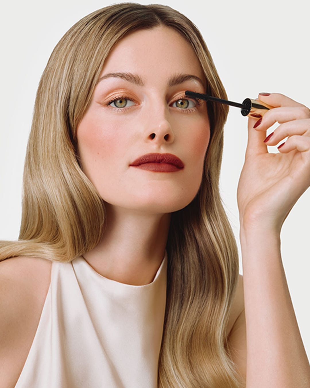 Eine Frau mit blondem Haar und rotem Lippenstift trägt Mascara auf ihre Wimpern auf, wobei sie eine goldene Mascara-Tube mit schwarzem Bürstchen hält.