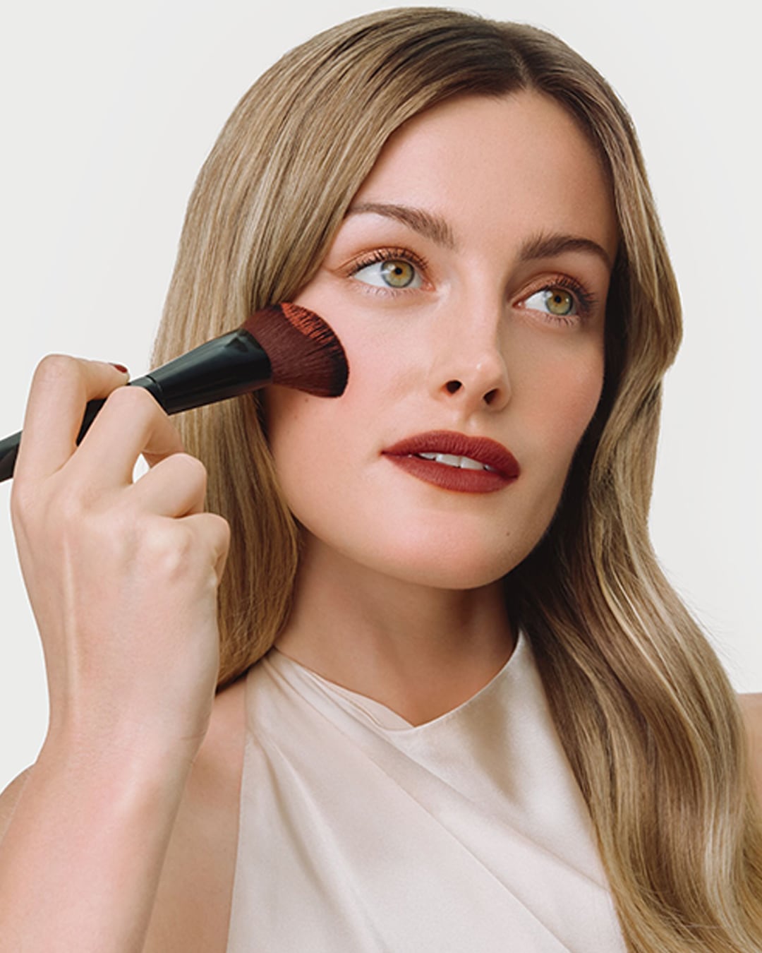 Eine Frau mit langen blonden Haaren trägt mit einem schwarzen Pinsel Rouge auf ihre Wange auf, während sie ein cremefarbenes Oberteil trägt und roten Lippenstift auf ihren Lippen hat.