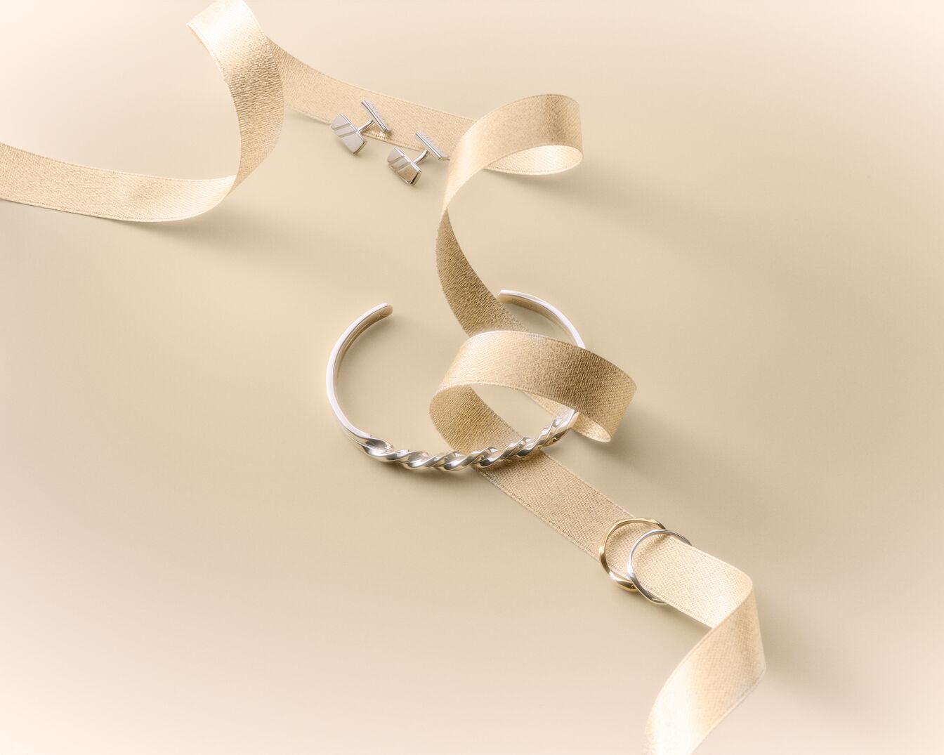 A silver twisted cuff bracelet, two silver cufflinks, and two rings, one silver and one gold, are elegantly displayed on a beige ribbon against a light beige background.