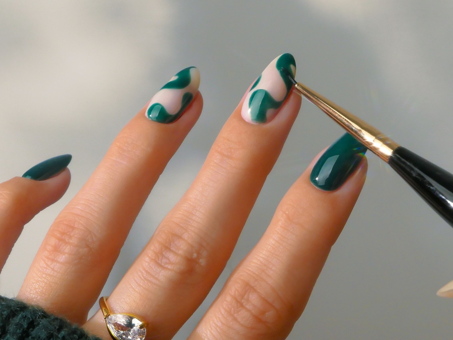 Eine Nahaufnahme zeigt eine Hand mit grün und cremefarben lackierten Nägeln, wobei ein Pinsel einen grünen Nagellack auf einen Nagel aufträgt, der ein abstraktes Muster erzeugt.