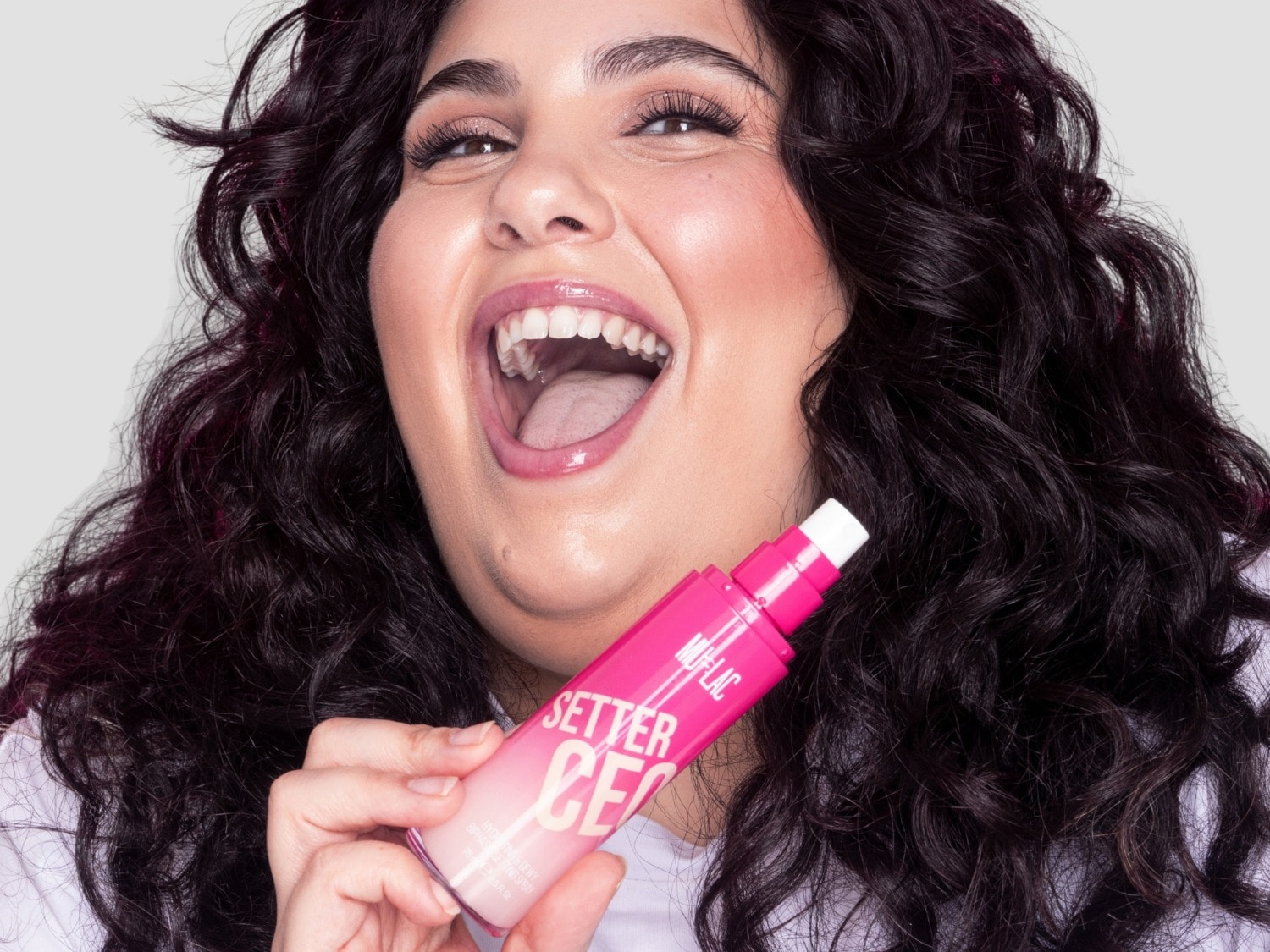 Una mujer sonriente con cabello oscuro y rizado sostiene un spray fijador de maquillaje rosa brillante de la marca "SETTER CEO" de MUA-LAC, con la boca abierta en una carcajada.