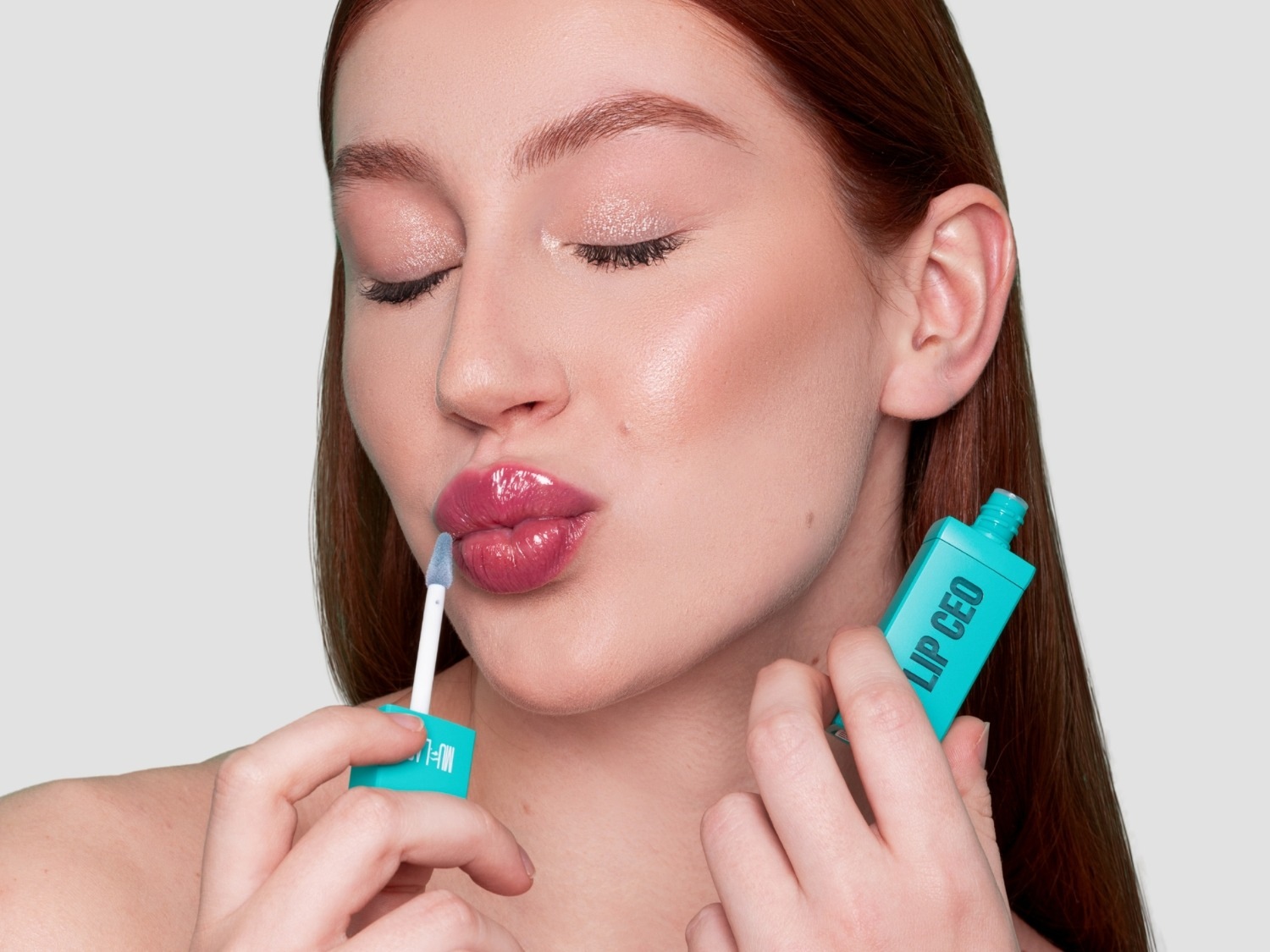 Una mujer con cabello castaño rojizo aplica un brillo labial rosa brillante de un tubo turquesa con la etiqueta "LIP CEO" en letras blancas, sus ojos están cerrados y sus labios ligeramente separados.