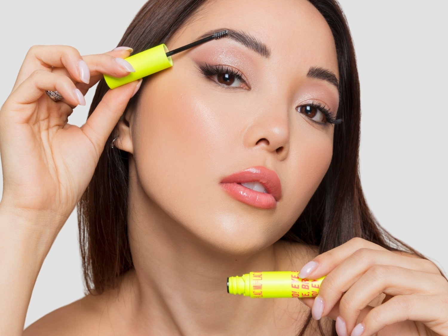 Una mujer con cabello castaño oscuro se aplica rímel en las cejas con un cepillo de un tubo de color amarillo neón, mientras sostiene la tapa del rímel en la otra mano.
