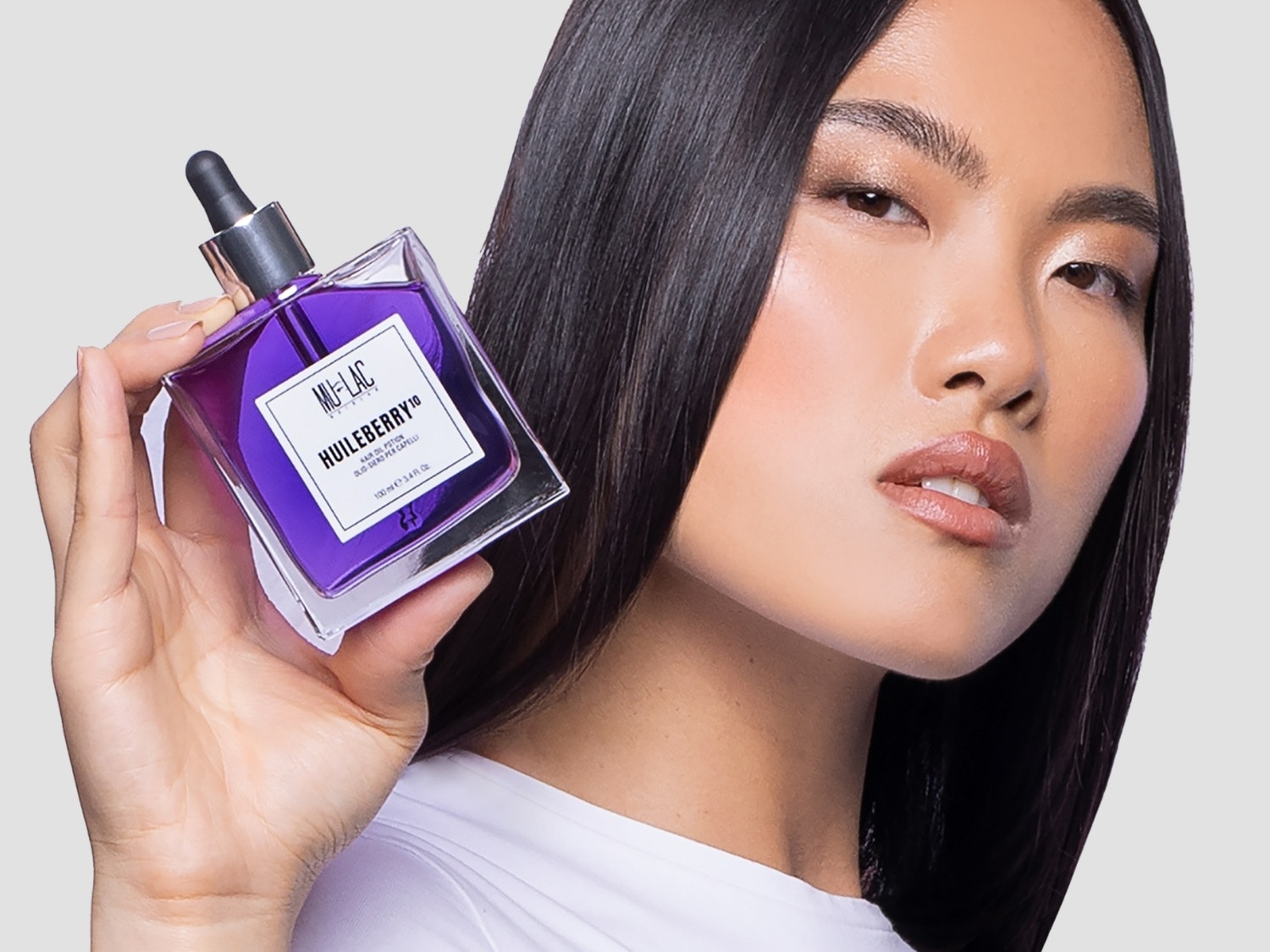 Una mujer con cabello oscuro y liso sostiene un frasco cuadrado de vidrio transparente con un gotero, lleno de un líquido violeta vibrante, con la etiqueta "MU-LAC HUILEBERRY 10" en blanco.