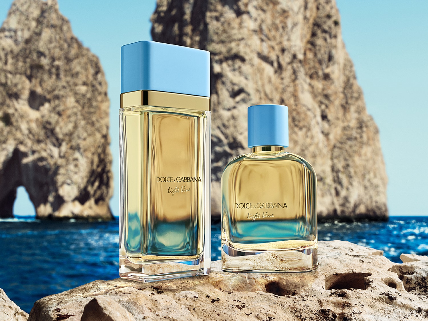 Deux flacons de parfum Dolce & Gabbana Light Blue, l'un rectangulaire et l'autre arrondi, avec des capuchons bleu clair et un liquide doré, sont posés sur un rocher texturé au bord de l'eau, avec des formations rocheuses en arrière-plan.