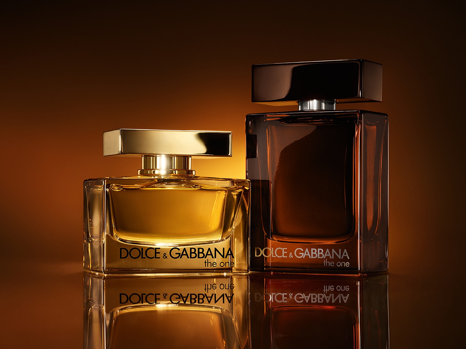Deux flacons de parfum Dolce & Gabbana The One, l'un doré et l'autre ambré foncé, sont présentés sur une surface réfléchissante, avec un arrière-plan chaleureux et lumineux.