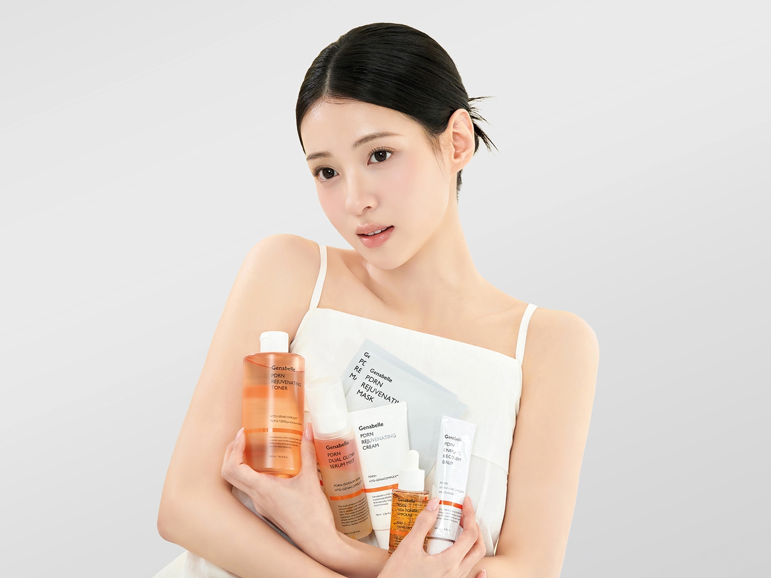Una donna con i capelli scuri raccolti all'indietro, indossa una canotta bianca e tiene in braccio una collezione di prodotti Genabelle PDRN Rejuvenating, tra cui un tonico, una maschera, una crema, un siero spray, una fiala e un balsamo recupero, tutti con etichette bianche e arancioni.
