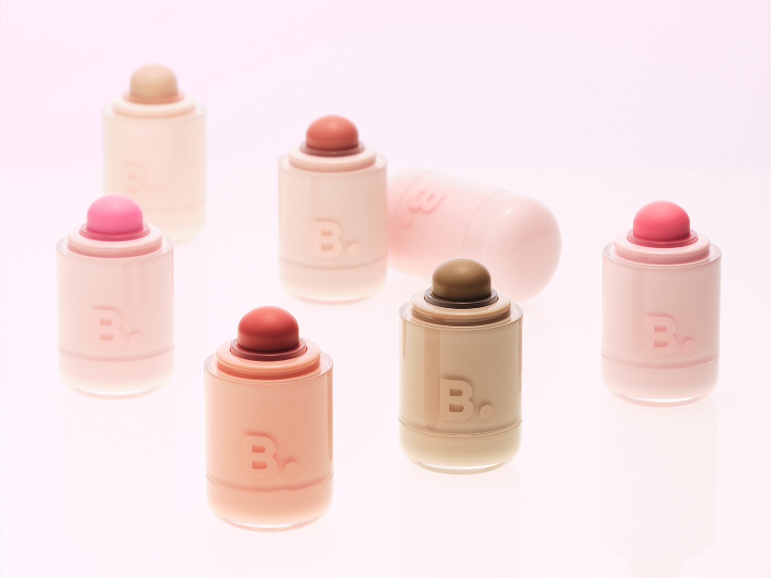 Sei stick di fard e illuminanti B. in tonalità pastello, tra cui rosa chiaro, pesca, marrone e beige, sono disposti su uno sfondo rosa chiaro, con uno stick rosa chiaro sdraiato orizzontalmente sullo sfondo.