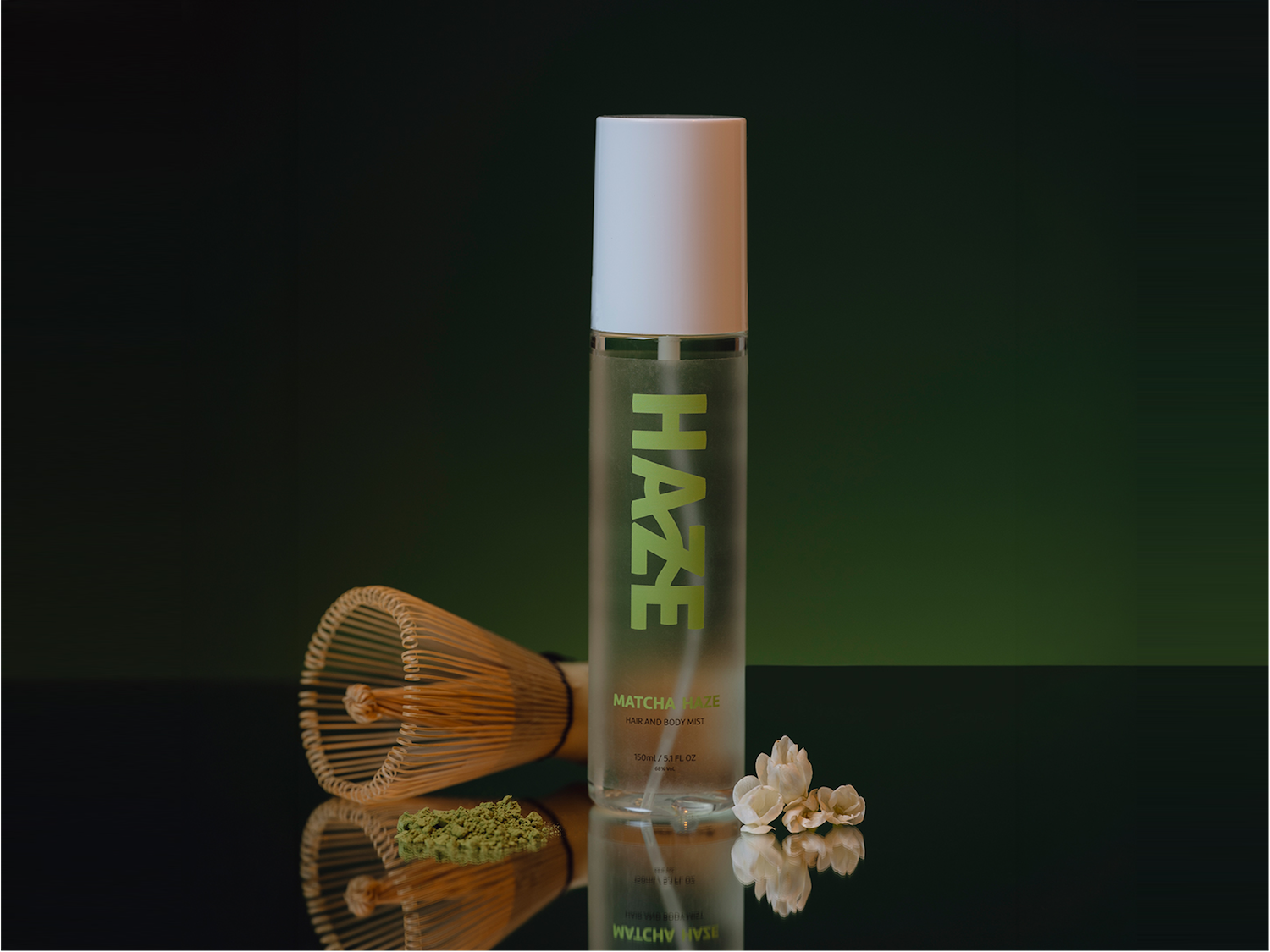 Le flacon transparent de brume pour cheveux et corps Matcha Haze de 150 ml, avec son bouchon blanc, est posé à côté d'un fouet en bambou et d'une pincée de poudre de matcha, le tout reflété sur une surface sombre.