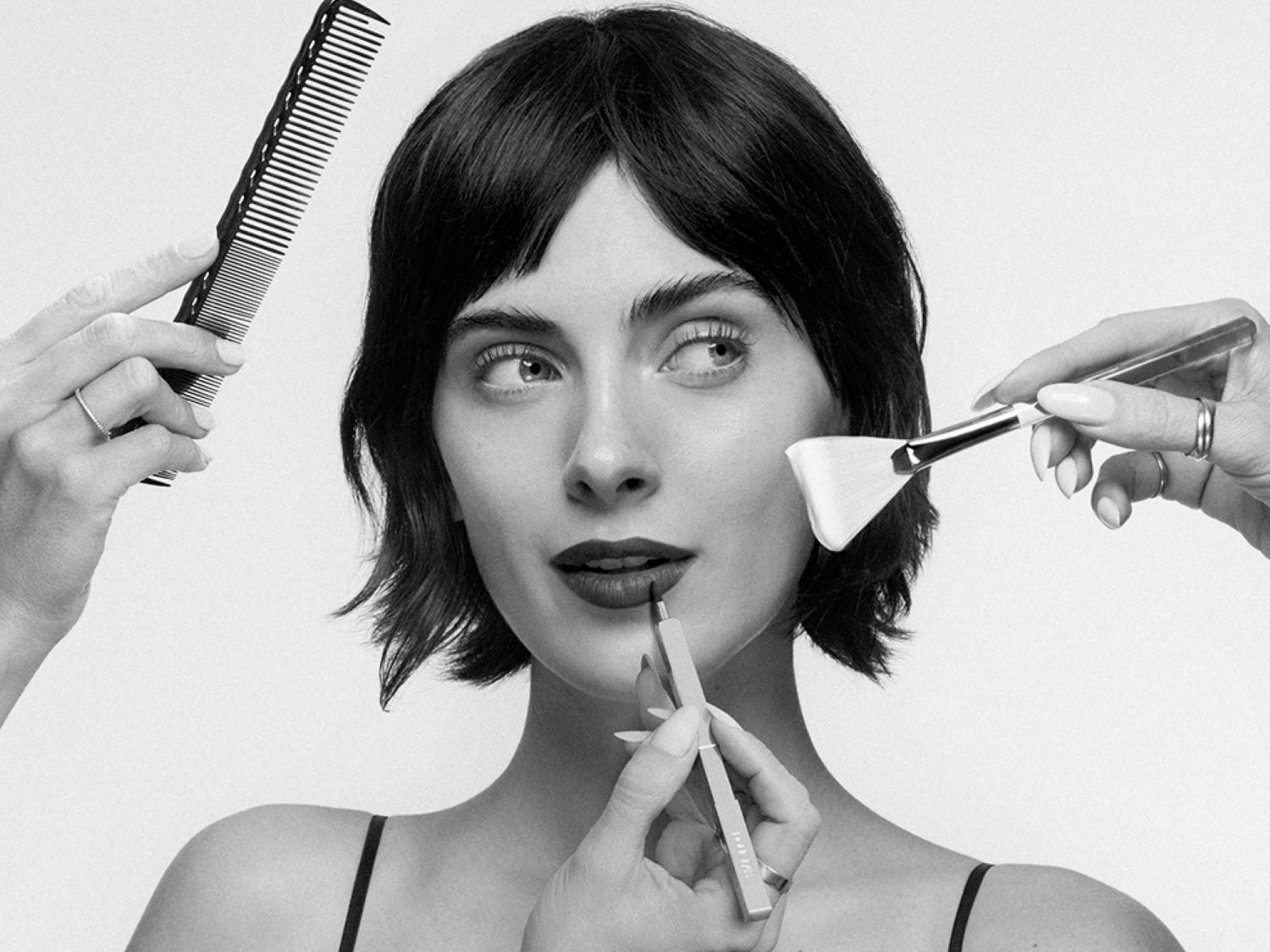 Una donna con capelli corti e scuri, labbra rosse, è circondata da mani che applicano trucco e acconciano i capelli, con un pettine nero a sinistra, un pennello per il viso a destra e una matita per labbra in basso.