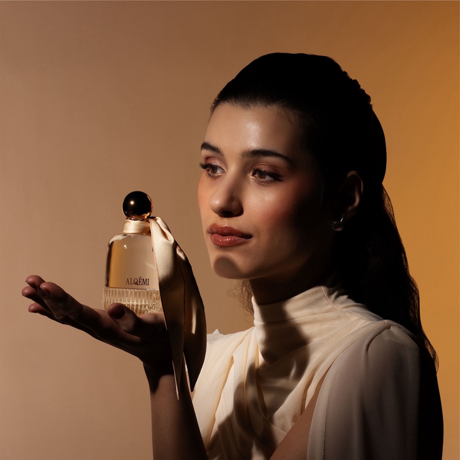 O femeie cu părul închis la culoare, cu o bluză crem, ține în palma deschisă un parfum ALQEMI, cu o fundă crem legată la gâtul sticlei, pe un fundal maro deschis.