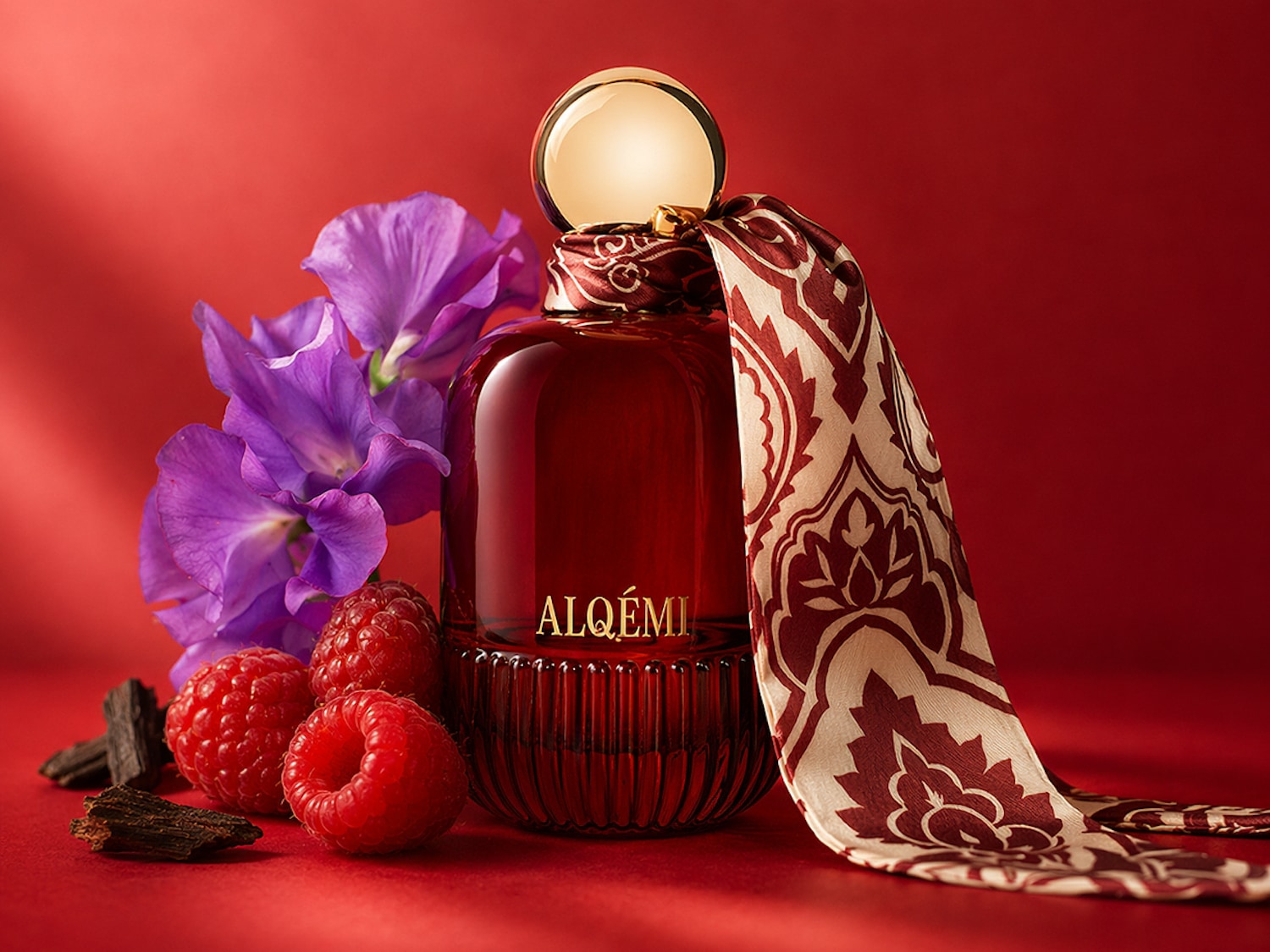 O sticlă elegantă de parfum Alqemi de culoare roșu închis, cu un capac auriu, este așezată pe o suprafață roșie, înconjurată de zmeură proaspătă, flori violet delicate și bucăți de lemn, având o eșarfă cu model legată în jurul gâtului sticlei.