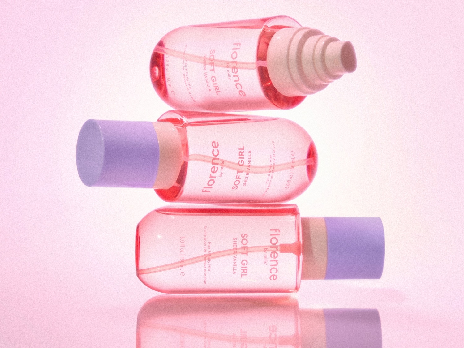 Three Florence by Mills Soft Girl Sheer Vanilla Hair & Body Mist bottles, each 5.0 fl oz / 150 ml, are stacked on a pink background, showcasing their light pink liquid and pastel purple caps.